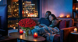 Couple cuddling on a sofa at night, watching TV with city lights outside and candles burning nearby.